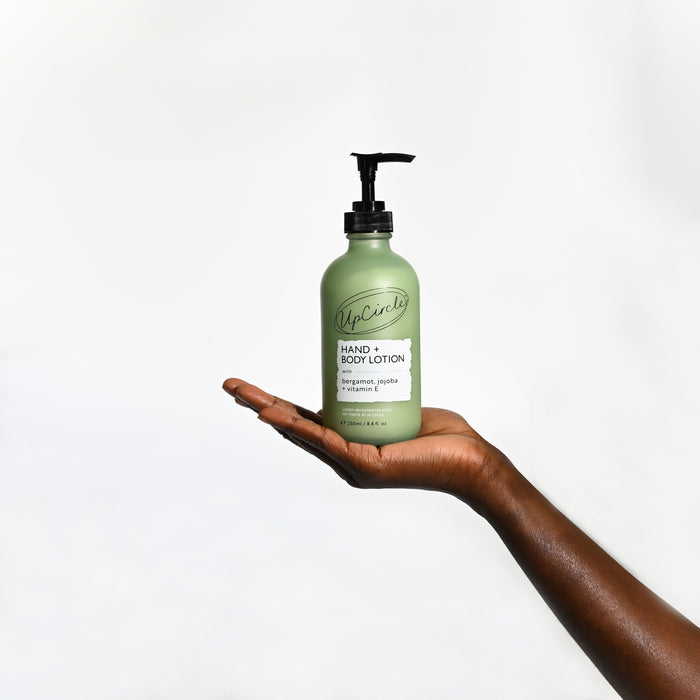 A hand holds a green pump bottle labeled UpCircle Hand + Body Lotion with Bergamot Water, a citrus-scented, natural body lotion infused with jojoba and shea butter. The background is plain white.