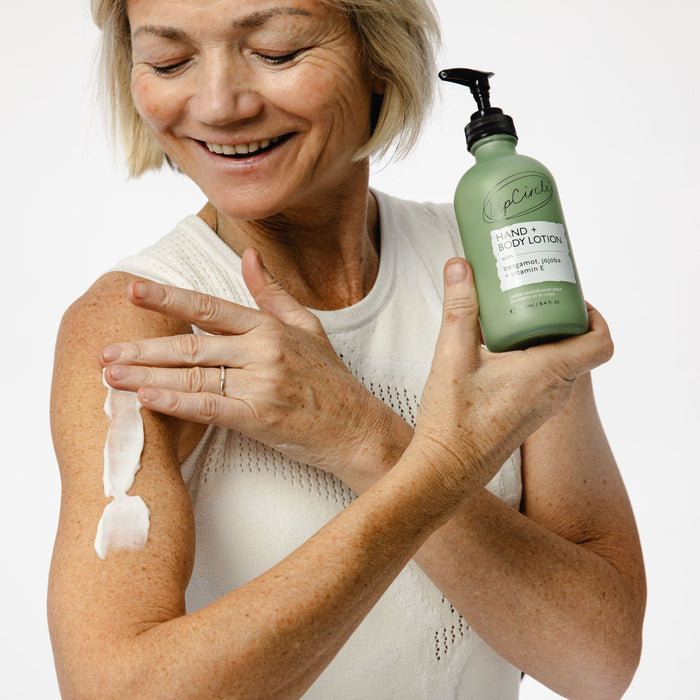 A person in a sleeveless white top applies UpCircles Hand + Body Lotion with Bergamot Water from a green bottle, smiling against a plain background.