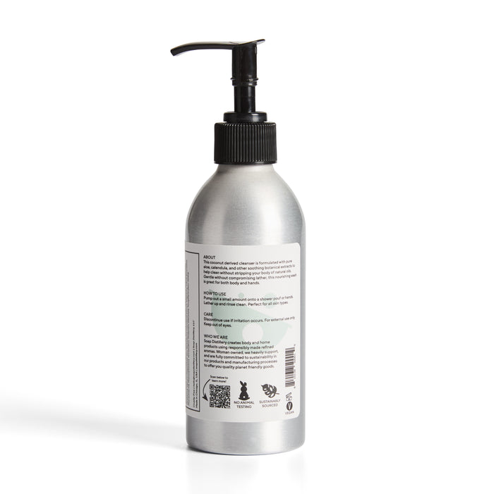 The Soap Distillery Hand + Body Wash comes in a silver aluminum bottle with a black reusable pump. It features sleek, minimalistic design elements on the label, including small text and icons, set against a plain white background.