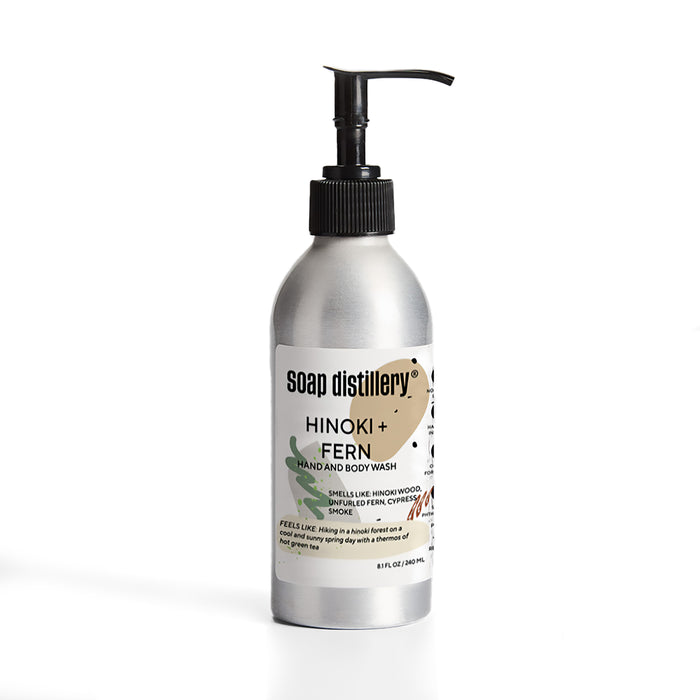 A silver aluminum bottle with a black pump labeled Soap Distillery Hand + Body Wash sits on a white background. The 8 fl oz (236 mL) label notes scents of hinoki wood, smoke, and fern.
