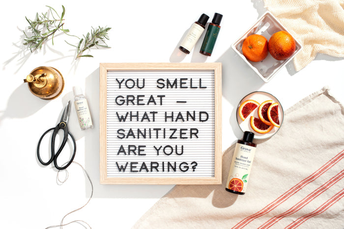 A Grove Co. letter board says, YOU SMELL GREAT - WHAT HAND SANITIZER ARE YOU WEARING? Surrounded by Grove Co. Hand Sanitizer Gel bottles that kill 99.9% of germs, plus scissors, herbs, oranges, and a striped cloth on a white surface.