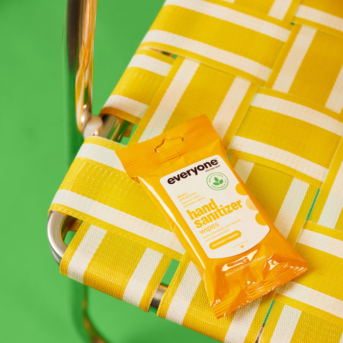 A pack of Everyone Hand Sanitizer Wipes, featuring refreshing lemon and coconut scents, rests on a yellow and white woven chair against a green backdrop. The orange pack, designed with white text and graphics, complements the vibrant scene beautifully.