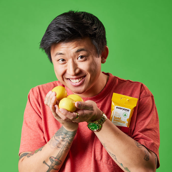 A smiling person in a red shirt holds two lemons and has an Everyone Hand Sanitizer Wipes packet tucked in their pocket against a bright green background.