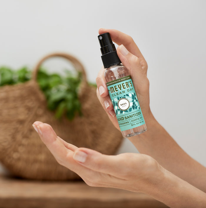 A person holds a bottle of Mrs. Meyers Hand Sanitizer ready to spray onto their palm, and a woven basket is visible in the background.