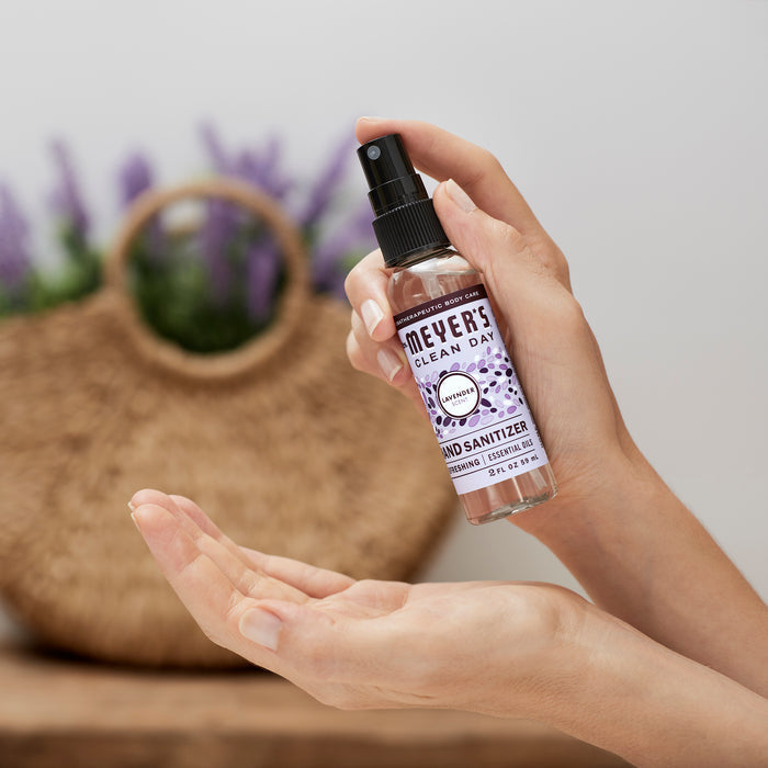 A person sprays Mrs. Meyers Hand Sanitizer with 62% ethyl alcohol into their palm, while a blurred woven bag in the background displays purple flowers, evoking cleanliness and nature.