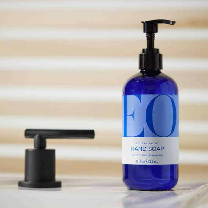 A blue EO Products hand soap bottle with a pump labeled Calming French Lavender for aromatherapy sits on a counter by a black faucet. The backdrop features blurred horizontal white and beige lines.