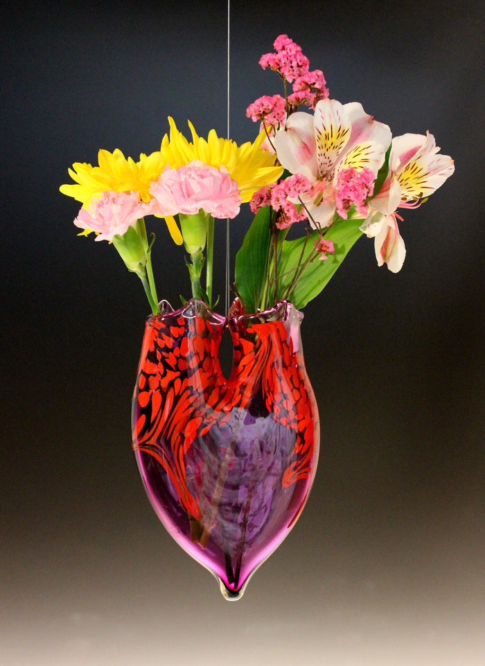 Hanging cremains vase in purple and red swirled glass holding yellow sunflowers pink carnations and white alstroemeria against a dark background