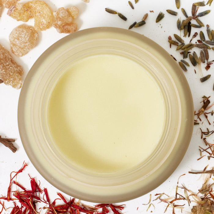 The Head to Toe Muscle Rub by WTHN sits on a white surface amid scattered herbs like lavender and saffron. It appears smooth and creamy, with a pale yellow hue, in a round jar.