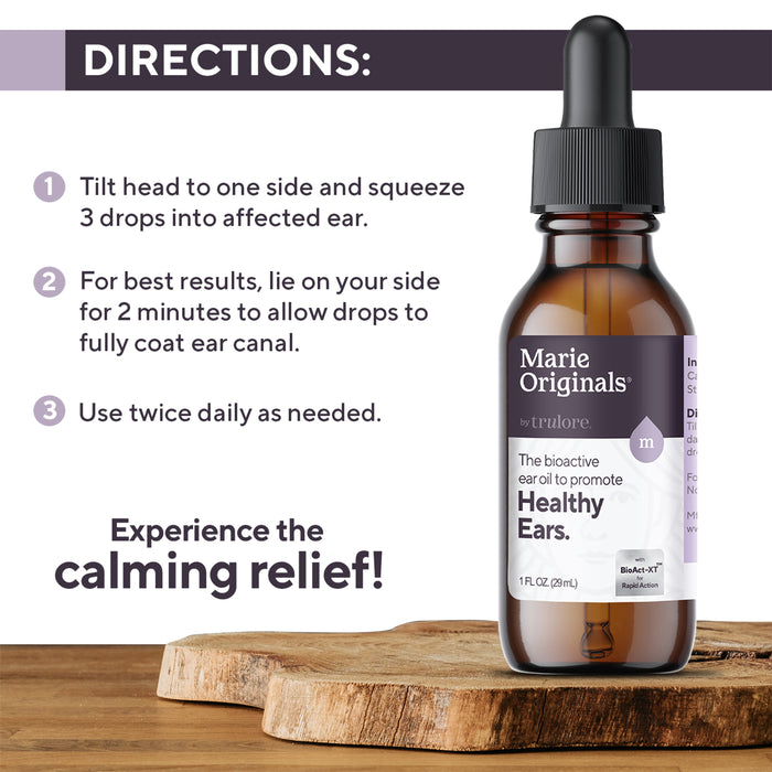 A bottle of Marie Originals Healthy Ear Oil with a dropper sits on a wooden surface. Instructions advise tilting your head, applying the oil, lying on your side, and using twice daily to support ear health.