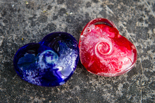 Two heart shaped glass cremains charms in blue and red on a textured rock surface.