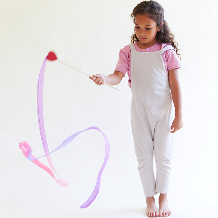 A young girl with curly hair, in a pink shirt and white overalls, barefoot on a light floor, uses Sarahs Silks Heart Streamer. She hones her gross motor skills as the streamers swirl in pink and purple patterns against a plain background.