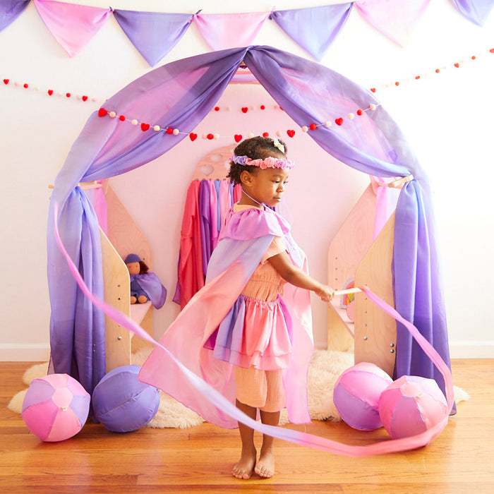 A child dances joyfully with Sarahs Silks Heart Streamer, showcasing gross motor skills in front of a play tent adorned with purple and pink fabrics. Two round cushions rest on the wooden floor, while festive red and white garlands hang above.