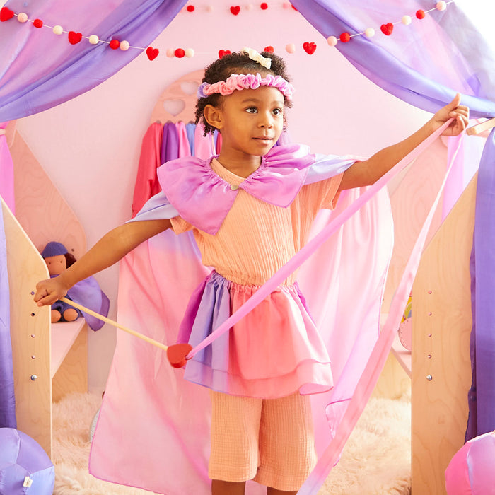 A child confidently stands between purple curtains in a pink and purple costume, complete with cape and headband. They hold a wand amidst heart garlands and Heart Streamers by Sarahs Silks, as toys eagerly await new adventures.