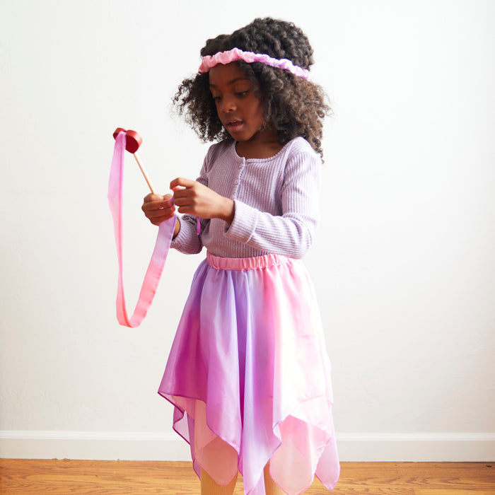 A young girl with curly hair is indoors, engaged in pretend play. Shes wearing a lavender top and pink-purple skirt, holding a Heart Streamer by Sarahs Silks to enhance her motor skills. Her matching headband and the wooden floor add simplicity to the scene.