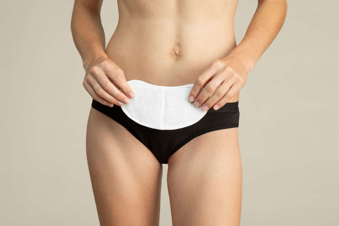 A person holds a large white pad against black underwear, focusing on the midsection with a beige backdrop, subtly highlighting the soothing power of Coras Heat Relief Patches infused with herbal extracts for menstrual cramp relief.