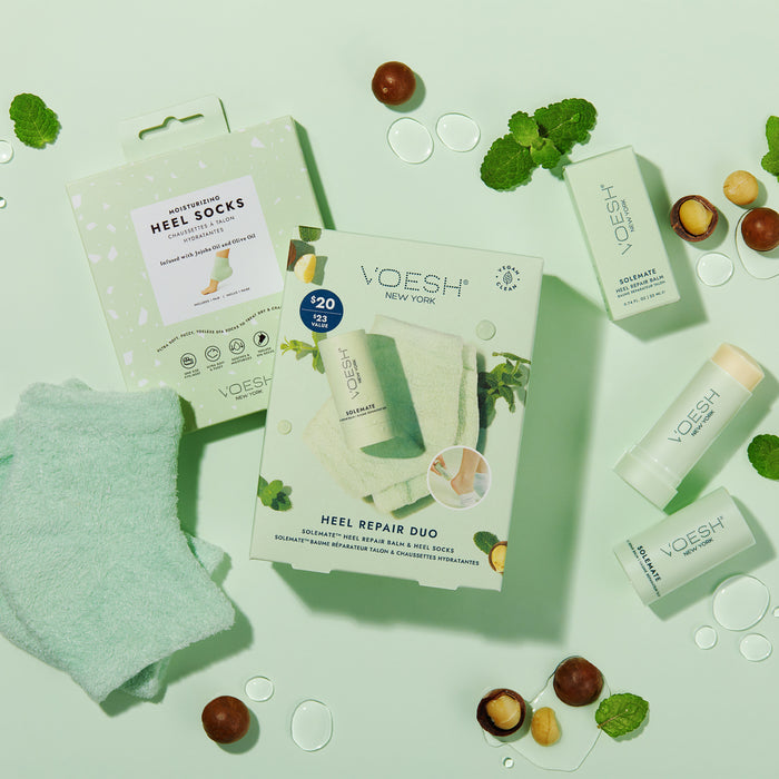 Flat lay of the VOESH New York Heel Repair Duo on a mint green background, featuring boxed moisturizing heel socks, Solemate Heel Repair Balm sticks, loose almond and mint leaves, and spherical droplets for a fresh touch.