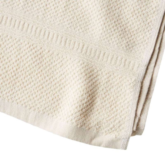 Close-up of a textured, off-white Hemp and Organic Cotton Bath Towel by Anact, showcasing a decorative border. Its surface displays a subtle waffle-like pattern for enhanced softness, with neatly hemmed edges that highlight craftsmanship.