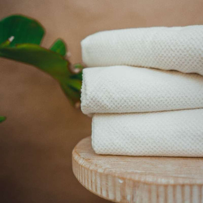 Three neatly folded Anact Hemp and Organic Cotton Bath Towels rest on a wooden surface, with a green plant in the background, conveying tranquility and cleanliness.