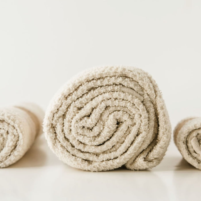 A neatly rolled beige towel from Anacts Hemp and Organic Cotton Bath Towel Set is centered, with parts of two others visible. The simple, light background creates a calm atmosphere that highlights the natural beauty of the organic cotton.