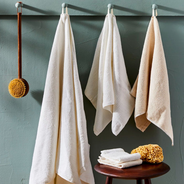 Three Anact light-colored, dye-free towels hang on hooks against a muted green wall with a wooden bath brush. Below, a small wooden table holds folded Hemp and Organic Cotton Bath Towels and yellow natural sponges.