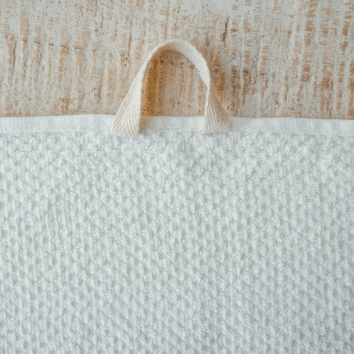 An Anact Hemp and Organic Cotton Hand Towel, in textured white, elegantly hangs on a rustic wooden surface. The loop at the top center contrasts with the woods rough texture.