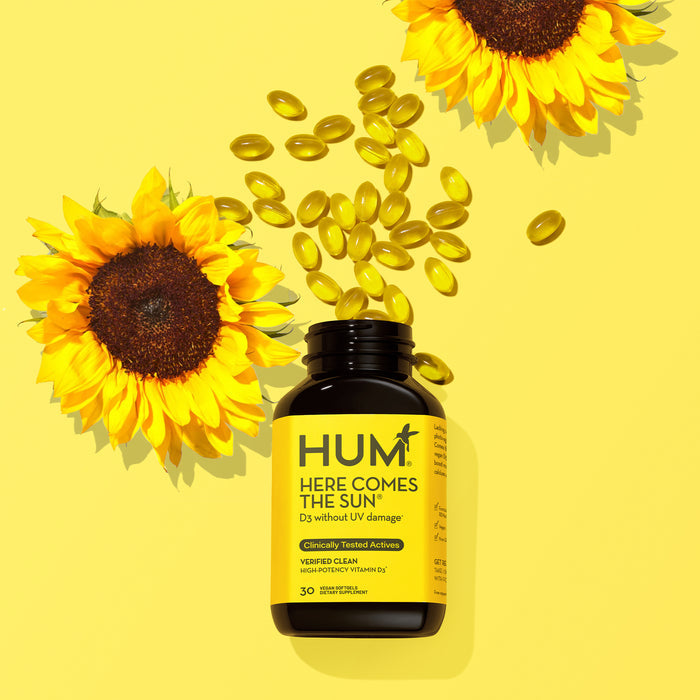 A sleek black bottle of HUM Nutritions Here Comes The Sun Vitamin D3 Supplement, with vibrant yellow labels, spills capsules next to a bright sunflower on a yellow background, promising immune support.