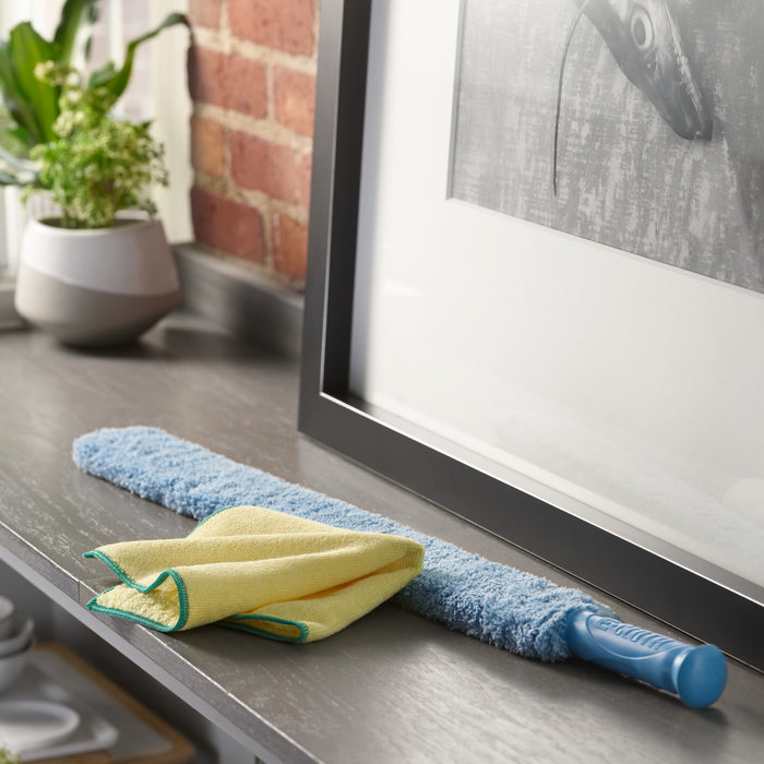 A long blue duster and an E-Cloth High Performance yellow microfiber cloth sit on a gray shelf, which is adorned with a small potted plant. A framed black-and-white photo leans against the exposed brick wall.