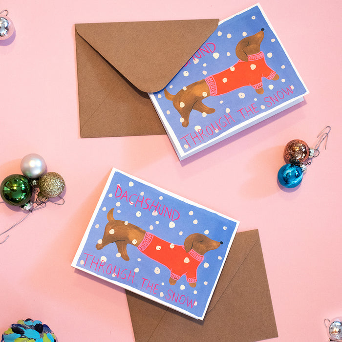 Two Holiday Cards - Dog by Supra Endura featuring a dachshund in a red sweater with Dachshund Through the Snow sit on a pink surface. Great for dog lovers, both cards and envelopes use recycled paper. Colorful ornaments and hooks are nearby.