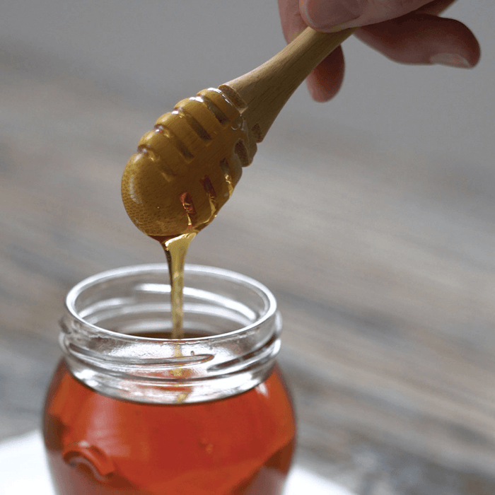 A hand holds a Bambu Honey Dipper, letting golden honey flow into a clear glass jar on a wooden surface.