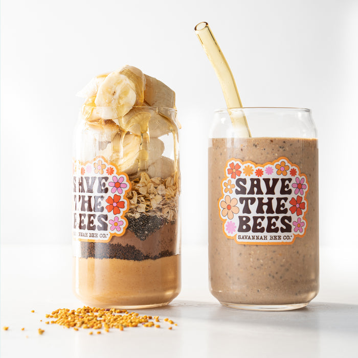Two jars reading Save the Bees sit next to each other. One contains layers of peanut butter, chia seeds, granola, and banana slices. The other features a brown smoothie with a Savannah Bee Company Honey Straw poking out. Bee pollen is sprinkled in front for added sweetness.