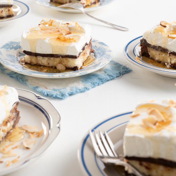Layered dessert slices with cream, chocolate, and nuts rest on decorative plates topped with caramel drizzle and almond slices. On the side are BPA-free Savannah Bee Company Honey Straws filled with acacia honey on a white surface for added sweetness.
