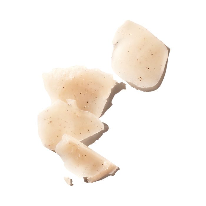 Several irregular, translucent, pale beige shards with small brown specks—reminiscent of Ursa Major’s Morning Mojo Bar Soap—are scattered on a white background, emphasizing the exfoliating qualities of this natural cleansing bar.