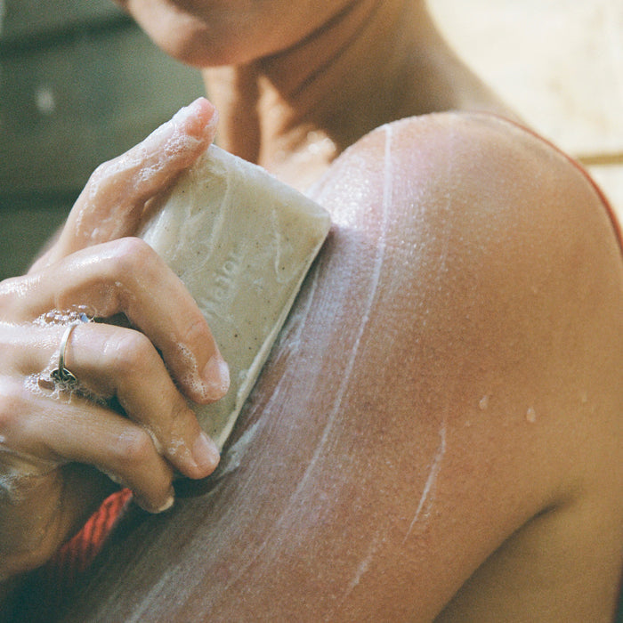 A person in a red top lathers their shoulder with Ursa Major Morning Mojo Bar Soap, producing foam and suds as the natural bar gently exfoliates skin while highlighting their hand, soap, and shoulder.