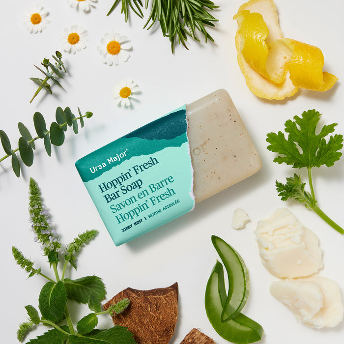 A bar of Ursa Major Morning Mojo Bar Soap, featuring natural ingredients like citrus peel, chamomile flowers, aloe, and shea butter, lies unwrapped beside fresh herbs. Its turquoise packaging with white and black text rests on a white background.