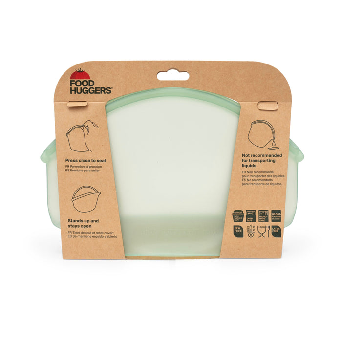 A green Hugger Bag by Food Huggers rests in cardboard packaging with use instructions and illustrations. Its versatile, easy to clean, dishwasher safe, and microwave safe, simplifying meal prep.