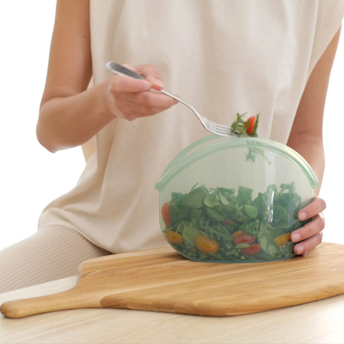 A person in a beige top holds a fork and a Hugger Bag from Food Huggers filled with a colorful salad. The vibrant greens, cherry tomatoes, and sliced veggies enhance their wooden cutting board seating as they enjoy the microwave-safe ease of the container.