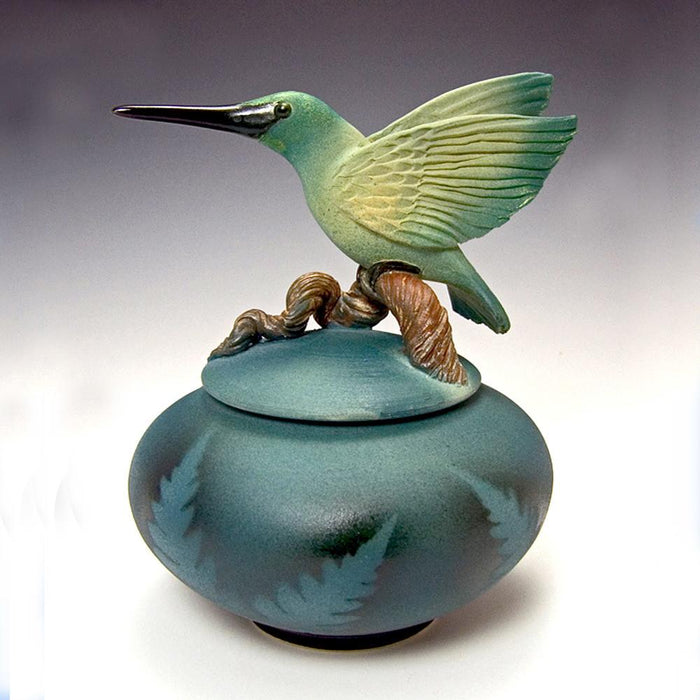 Handmade ceramic urn jar for ashes featuring a green hummingbird perched on a branch atop a blue round vessel.