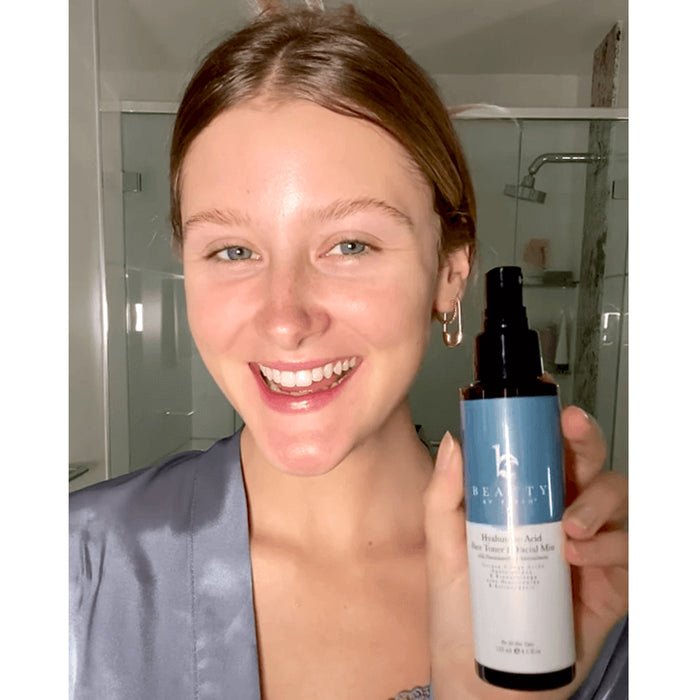 A light-haired person in a blue top smiles in the bathroom while holding a bottle labeled Hyaluronic Acid Toner by Beauty by Earth, essential for moisture retention and achieving a youthful complexion.