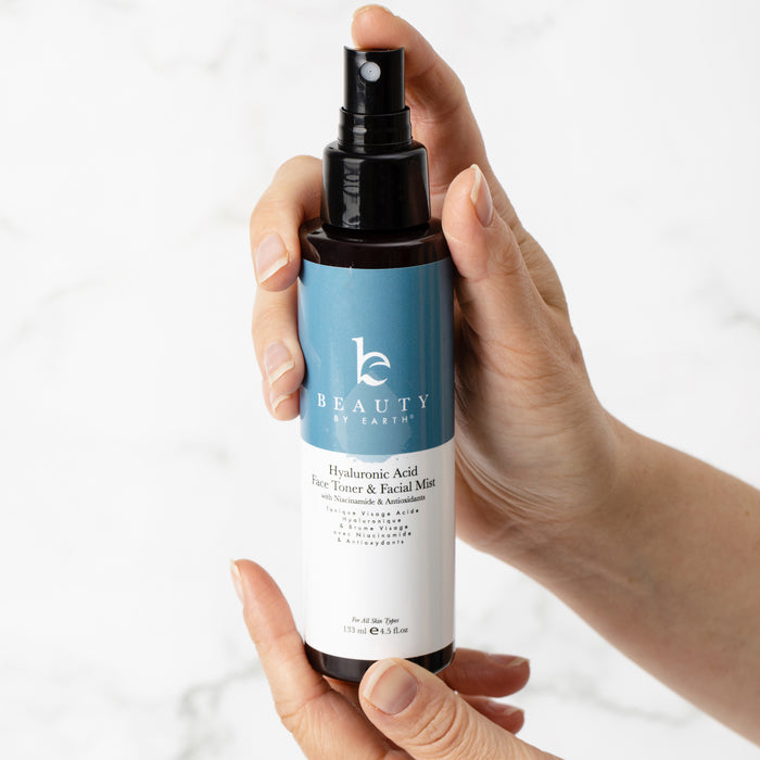 Hands hold a Beauty by Earth Hyaluronic Acid Toner with a blue and white label, featuring a black spray nozzle. Set against a light marbled surface, its ideal for moisture retention and promises to enhance youthful complexion.