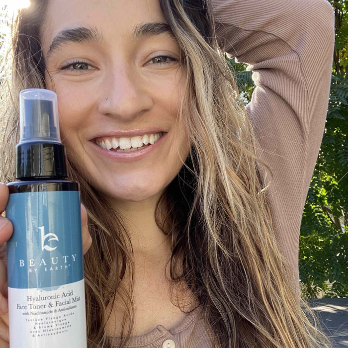 A woman with long hair stands outdoors, smiling radiantly as she holds a bottle of Beauty by Earth Hyaluronic Acid Toner. Surrounded by greenery, her glowing complexion suggests excellent moisture retention from the toner.