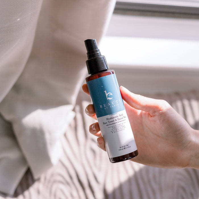 A hand holds a Beauty by Earth Hyaluronic Acid Toner in a dark amber spray bottle. Sunlight filters through, creating gentle patterns on the curtain behind it, while the stylish font promises moisture retention for youthful skin.