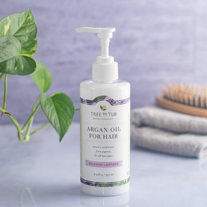 A bottle of Tree To Tub Hydrating Argan Oil Conditioner for Dry Hair & Scalp with a pump dispenser sits beside a green plant, brown hairbrush, and folded gray towels. Labeled Relaxing Lavender, this sulfate-free conditioner is perfect for dry hair, framed by a soft purple backdrop.