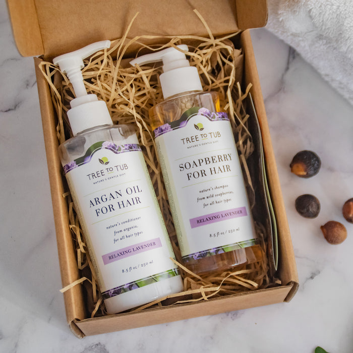 An open box on a marble surface holds two bottles nestled in straw. One reads Tree To Tub Argan Oil for Hair, the other Soapberry for Hair, both with a lavender scent. Nearby, scattered soapberries promise Tree To Tubs sulfate-free Hydrating Argan Oil Conditioner, perfect for rejuvenating dry hair and scalp.