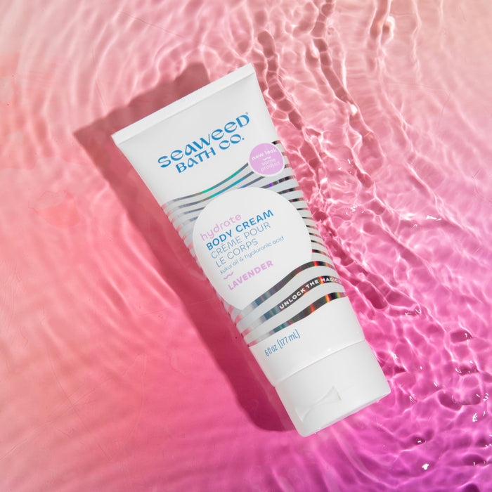 A tube of The Seaweed Bath Cos Hydrating Body Cream in Lavender scent rests on a pink, watery surface. The white packaging boasts blue text with stripe accents near the bottom, reflecting gently on the liquid.