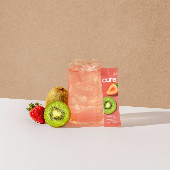 A glass of pink drink with ice sits beside a sliced and whole kiwi, a strawberry, and a Cure Hydration packet featuring kiwi and strawberry images on beige—delivering rapid absorption for dehydration relief with its Hydrating Electrolyte Drink Mix.