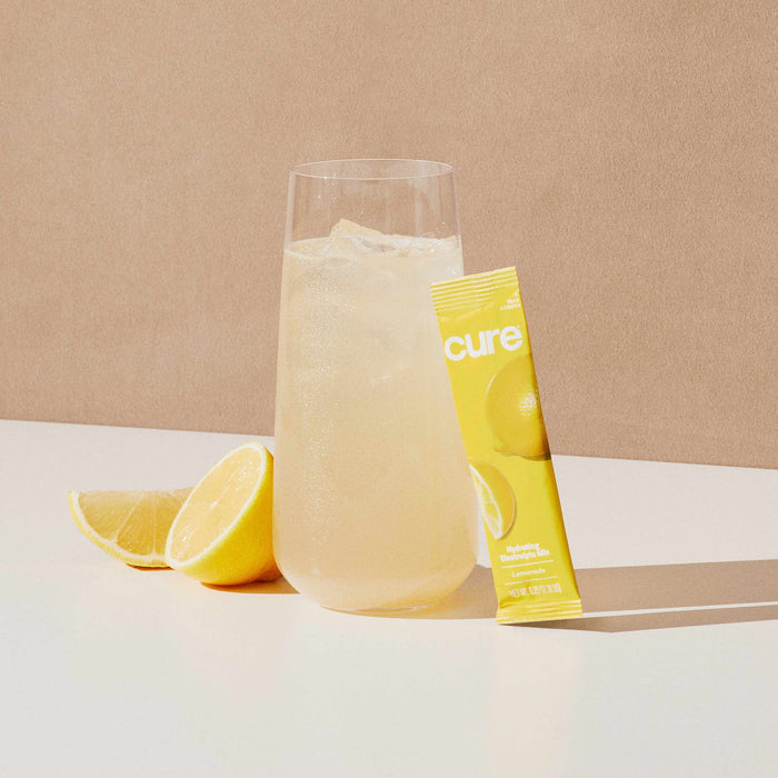 A tall glass of light-colored beverage with ice sits on a table next to sliced lemon and a yellow Cure Hydration packet adorned with lemon graphics, set against a neutral beige background, highlighting this refreshing Hydrating Electrolyte Drink Mix for rapid absorption.