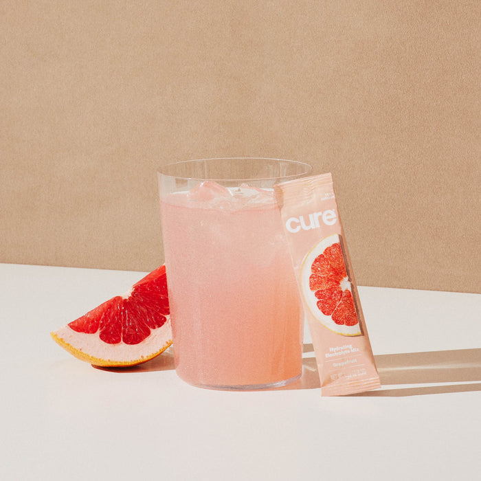 A glass of pink grapefruit drink with ice sits on a white surface, providing hydration relief. Next to it, a grapefruit slice and a Cure Hydration packet of Hydrating Electrolyte Drink Mix featuring a grapefruit illustration rest against a calming beige background.