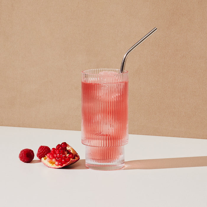 A tall, ribbed glass with pink Cure Hydration Electrolyte Drink Mix, ice, and a metal straw sits on a white surface. Surrounded by raspberries and an open pomegranate on a beige backdrop, this refreshing drink ensures rapid absorption for optimal hydration.