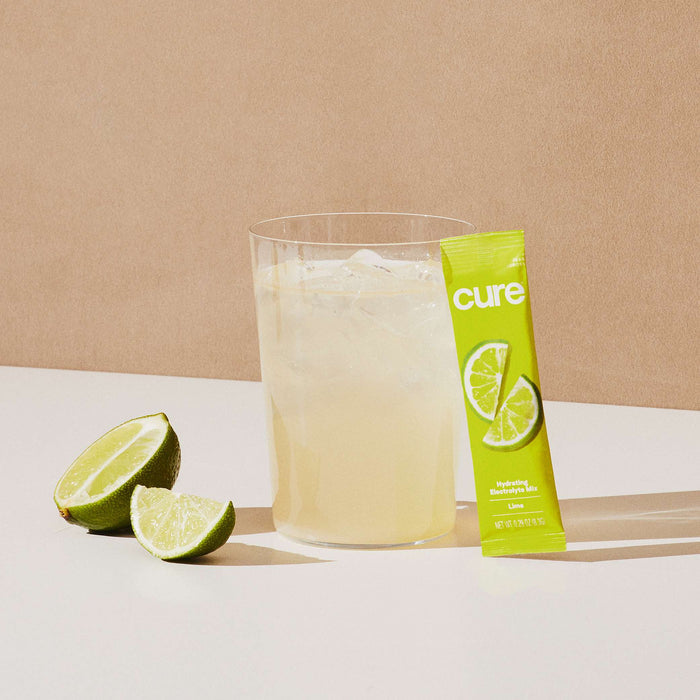 A glass of lemon-lime drink with ice is on a white surface, next to a bright green packet labeled Hydrating Electrolyte Drink Mix from Cure Hydration, a cut lime, and the promise of hydration akin to refreshing coconut water, set against a plain beige background.