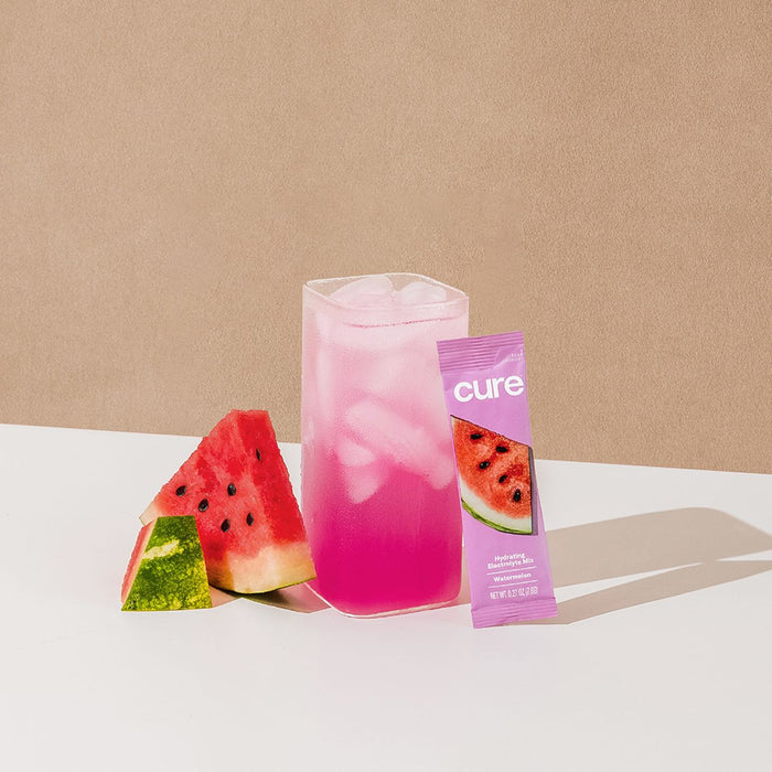 A tall glass of pink iced drink sits on a white surface, accompanied by two watermelon slices and a Cure Hydration packet. Promising rapid absorption and ultimate hydration, this Hydrating Electrolyte Drink Mix transforms into the perfect electrolyte experience against a beige background.